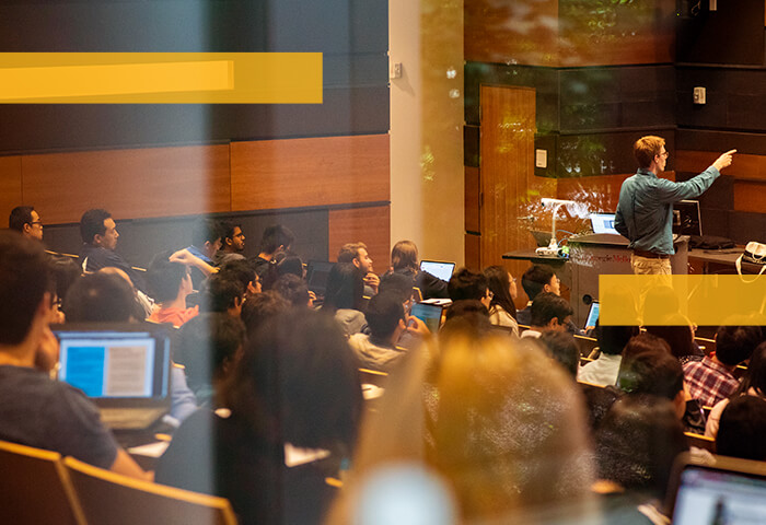 A lecture hall is filled with students listening to a professor speak at the head of the classroom.